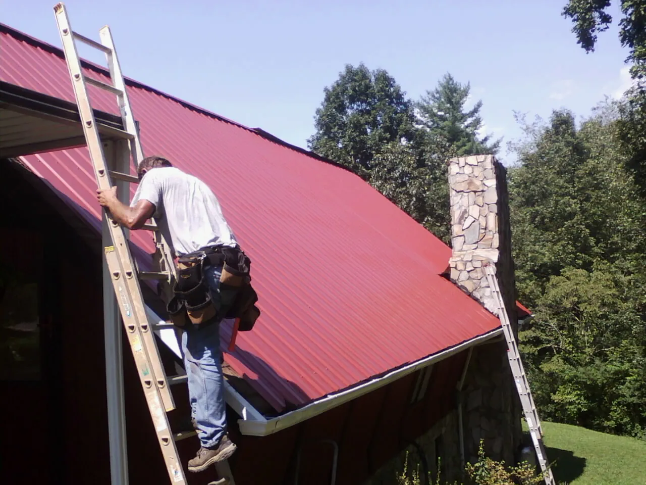 Roofer on a ladder beside a red metal roof and stone chimney — Soffit & Fascia Repair by Roofing Brothers in Somers Point